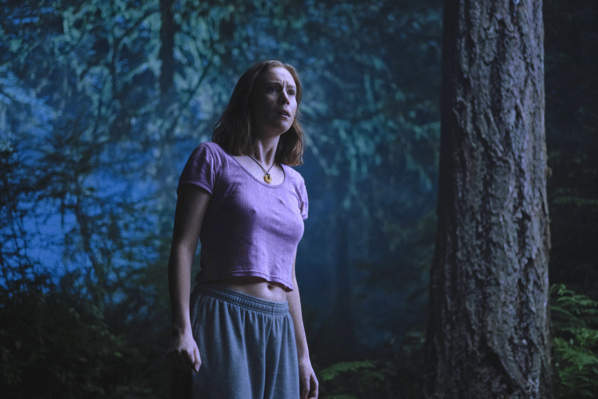 Hannah Einbinder as Kris wears a pink tee and sweatpants while standing in a forest at night in 'Teenage Sex and Death at Camp Miasma.'