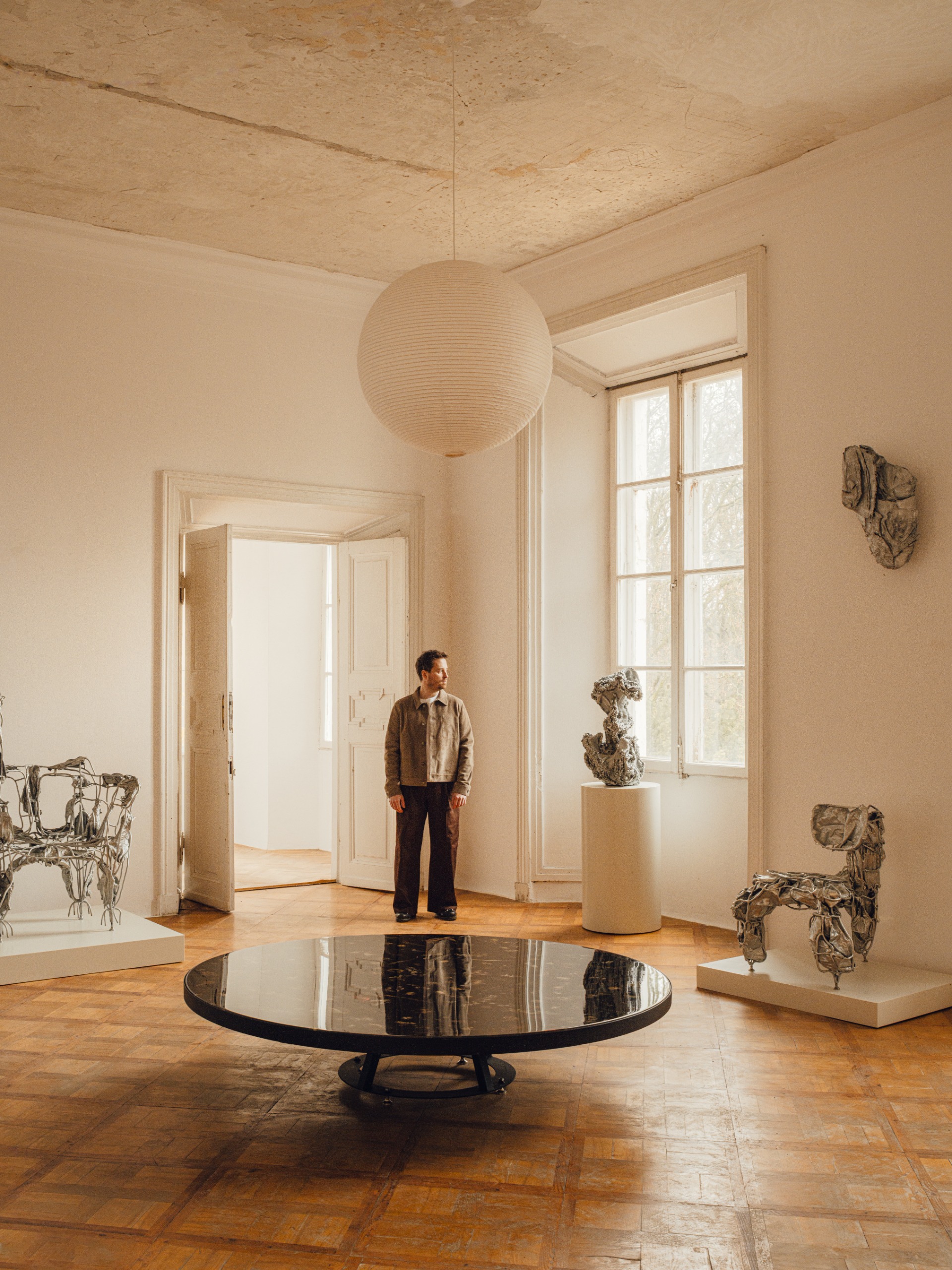 A young man in a gray overshirt and brown trousers stands in a naturally lit, palatial gallery filled with sculptural furniture in textured chrome and a resin round table looking outside a window.