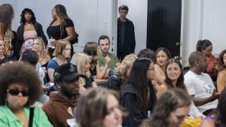 In the background, Nathan Fielder looks through a doorway into a room packed with casually dressed men and women, potentially about to audition to be in his latest epic "rehearsal" in The Rehearsal.