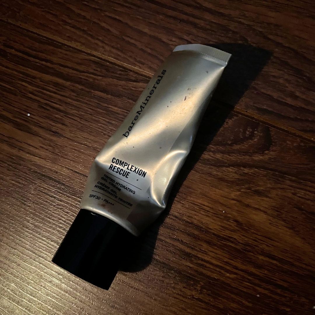 Empty gold tube of bareMinerals Complexion Rescue Tinted Moisturiser SPF30 with dark wood background