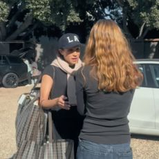 Meghan Markle wearing an LA baseball cap adn a black tee shirt carrying shopping bags and talking to a girl wearing jeans and a black tee with her backt o the camera in front of two cars, 