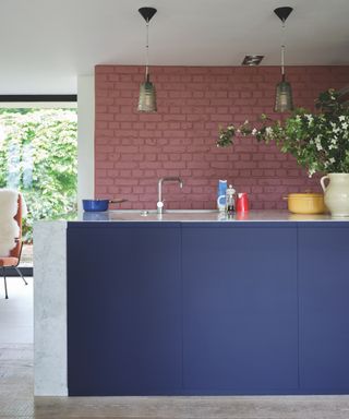 Pink and blue open plan kitchen with pink painted exposed brick walls, blue painted island, vase of spring blooms, pendant lights, lime washed floorboards