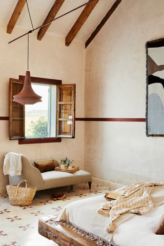 A rustic room with geometric tiled floor, a low wooden bed, red wooden shutters and a cream lounger