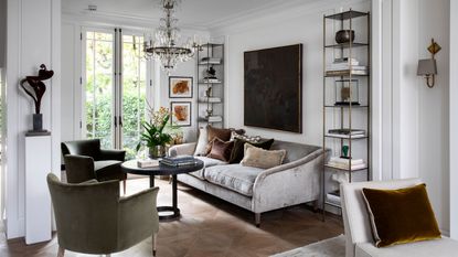Small living room storage ideas. Small living room with engaging seating area, large gray sofa facing two green lounge chairs, rounded black coffee table in the middle, two metal shelving units either side of the sofa, decorated with display items, large square artwork above sofa, dark painted color, low hanging traditional glass chandelier above coffee table, white painted paneled walls, warm wooden flooring 
