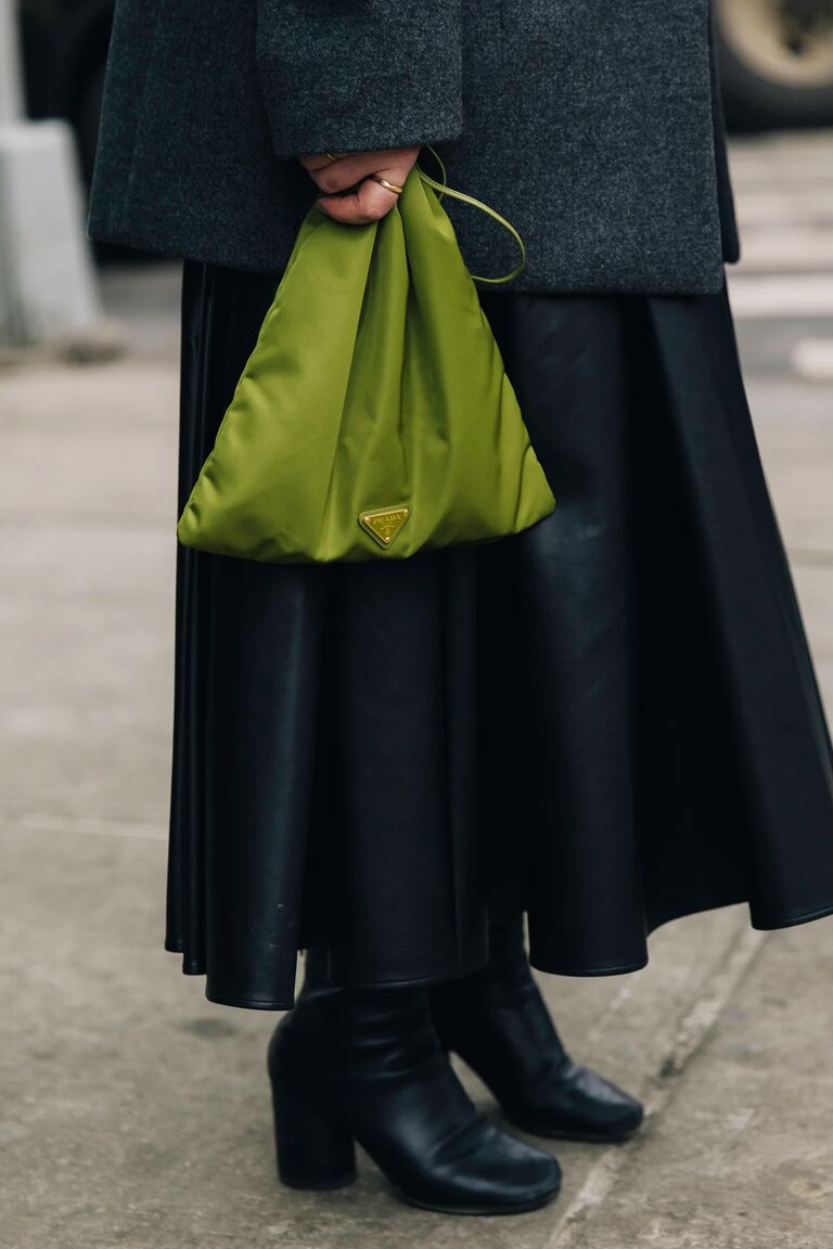 a woman at new york fashion week wearing a black skirt, black boots, and a green Prada pouch purse