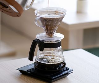A pour over coffee maker witha glass carafe on a wooden table with a gooseneck kettle pouring into it
