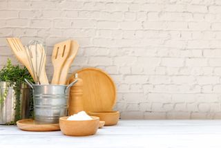 A white wall background behind a metal pot holding an array of wooden cooking utensils.