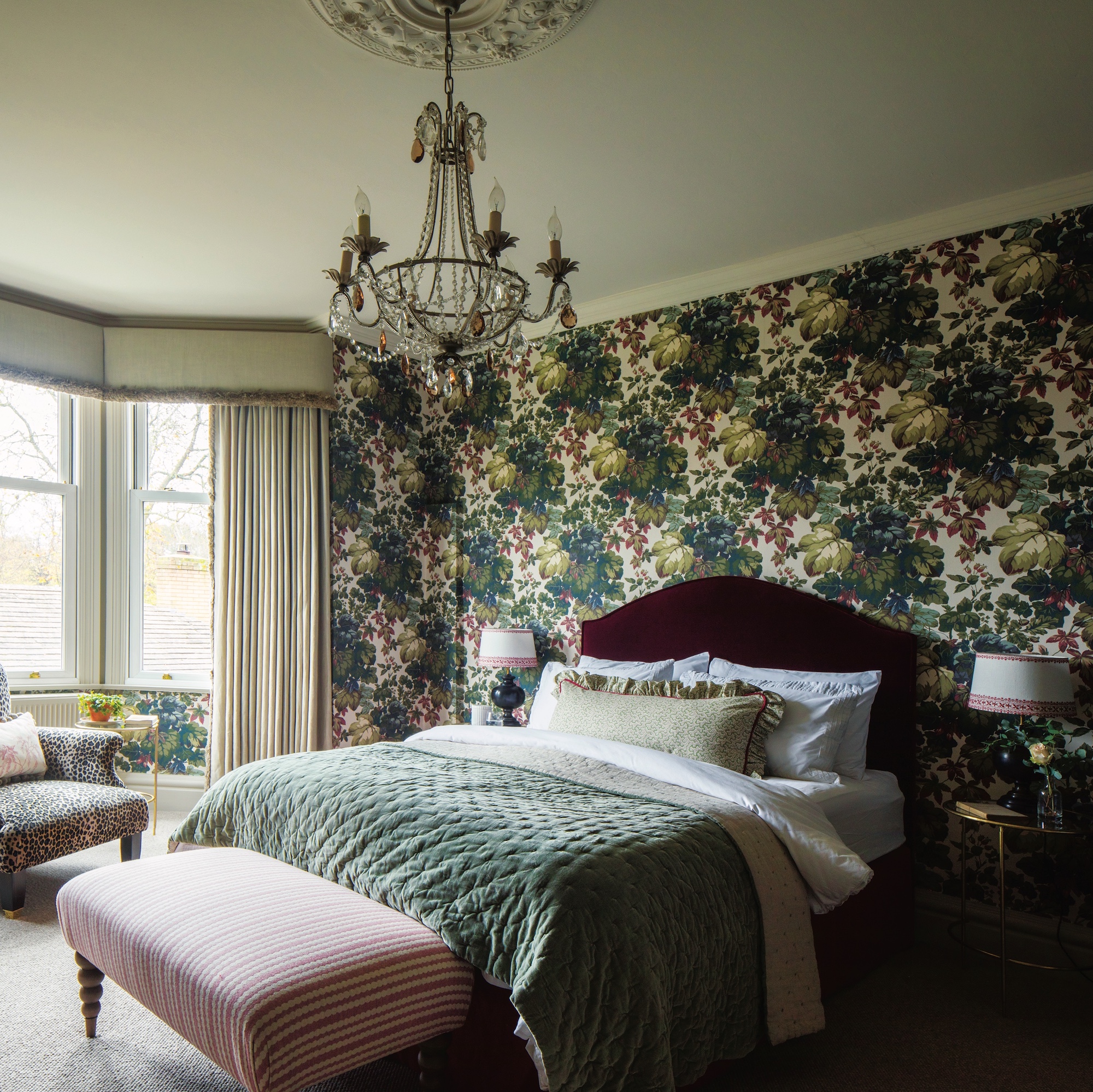 bedroom with dark botanical wallpaper in green and burgundy, and bed with burgundy headboard and green cover beneath a chandelier