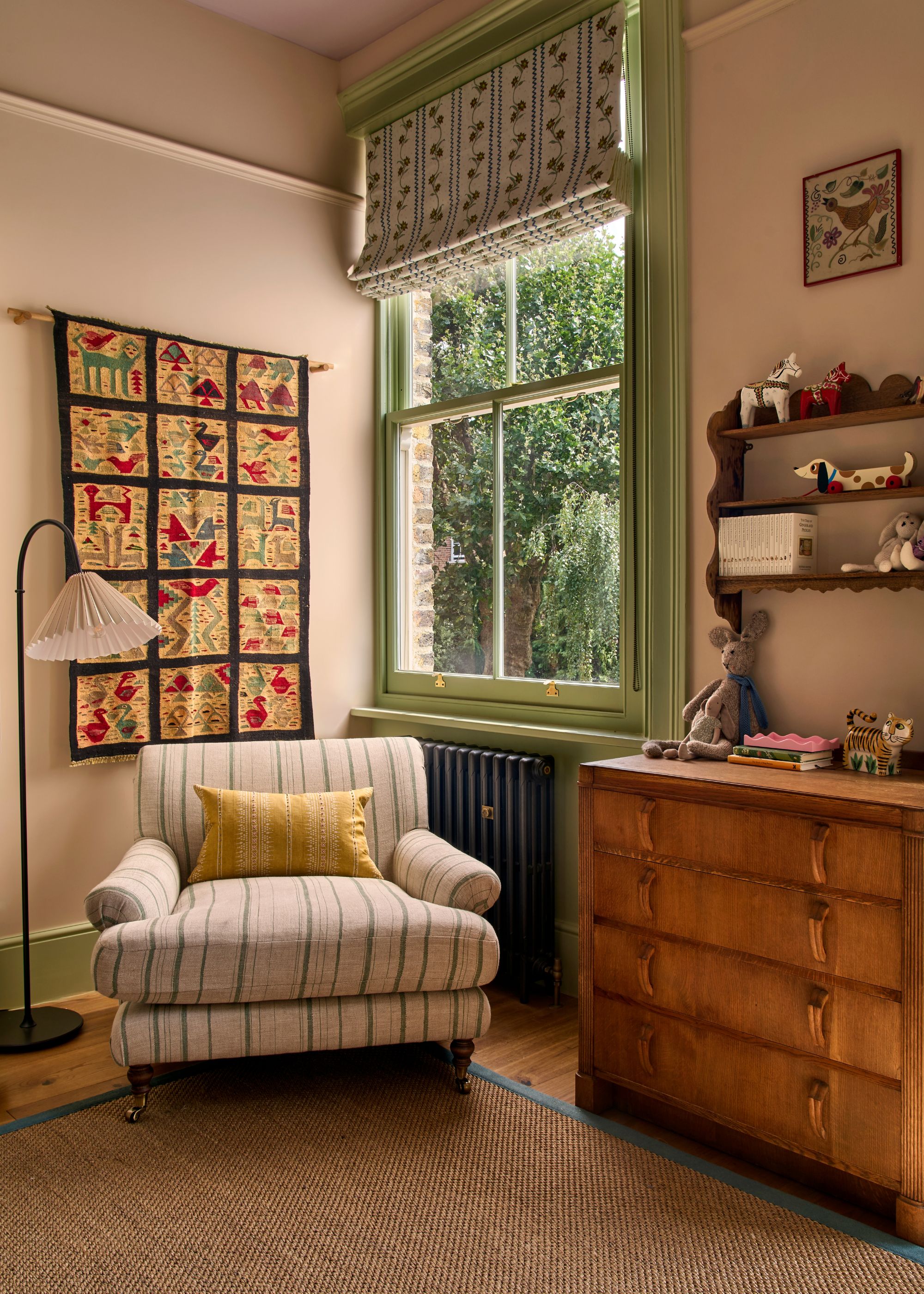 A kids' room with neutral walls, a sisal rug, and a striped armchair in the window, green window trim, patterned roman blinds, a wooden dresser, and a tapestry on the wall.