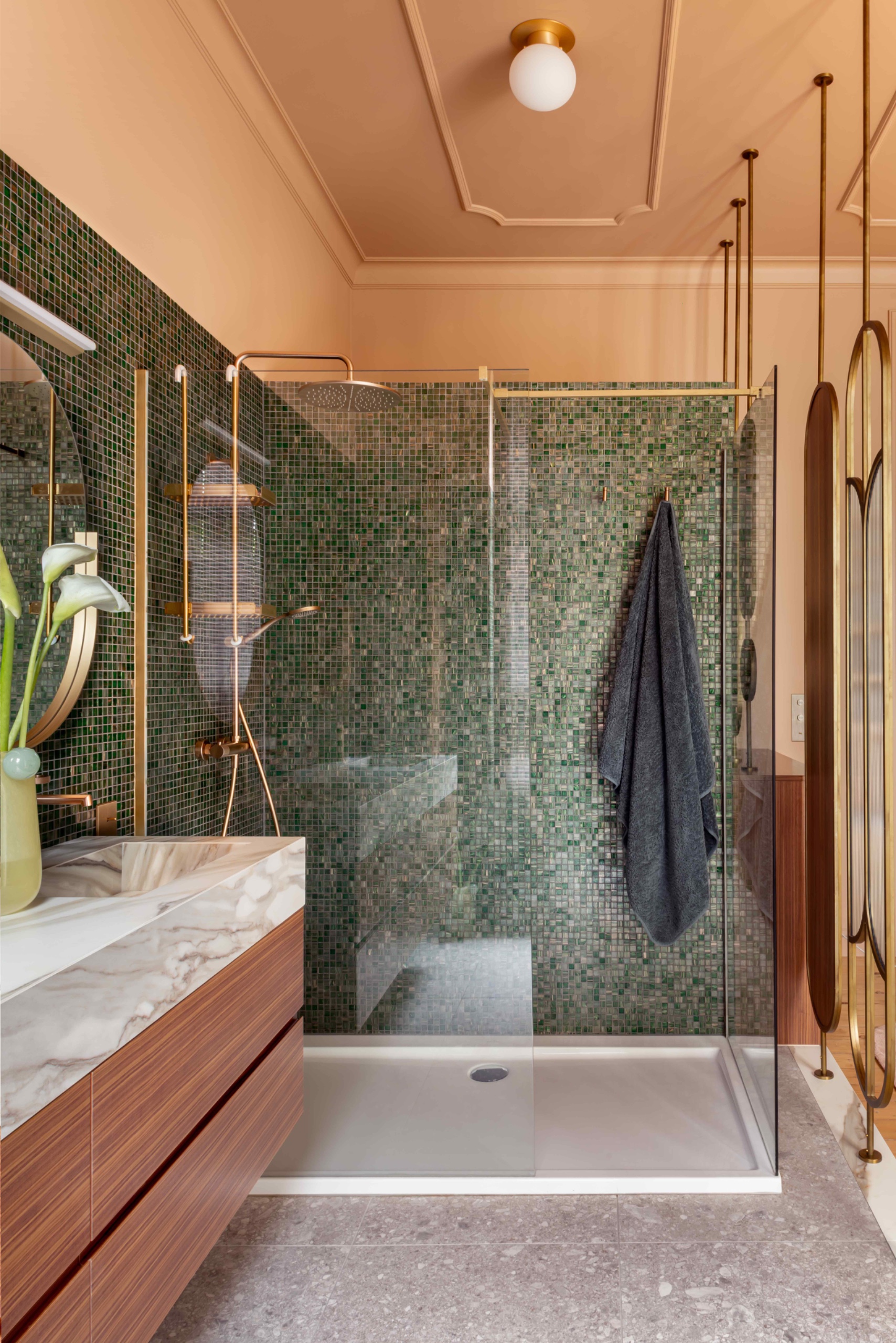 Bathroom with a slim white shower tray, green glass mosaic walls, brass fixtures, a floating wood vanity and a peach ceiling with decorative molding.