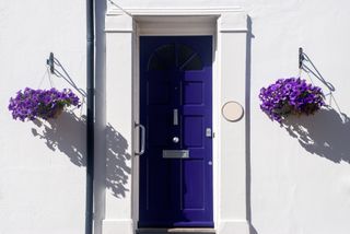 A white house with a blue front door flanked by two hanging baskets with purple petunia flowers