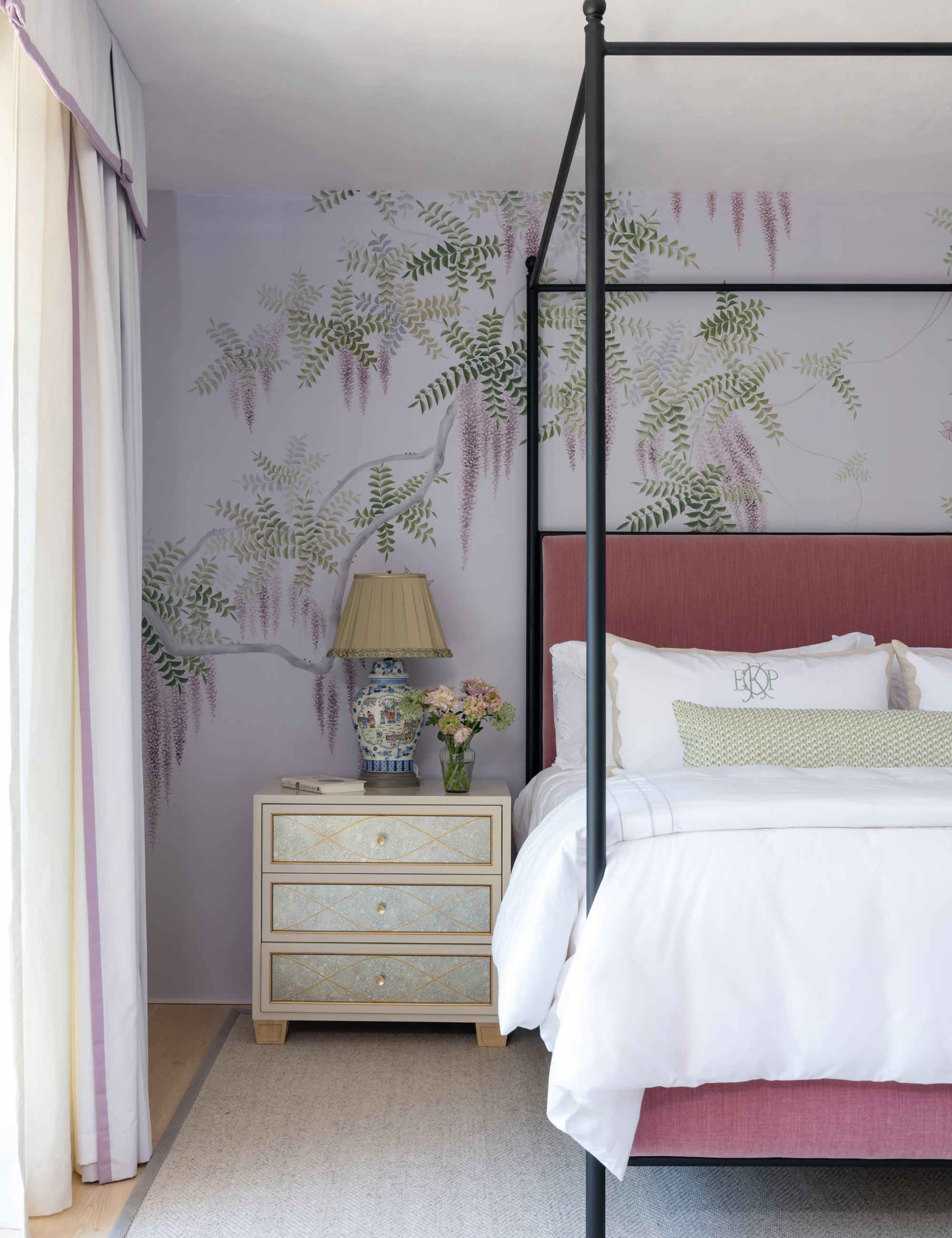 Summer bedroom featuring pale purple walls, an iron canopy bed, and a mirrored nightstand topped with a chinoiserie-style table lamp