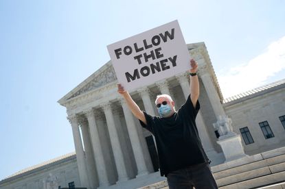 A man stands outside the Supreme Court with a sign that says Follow the Money