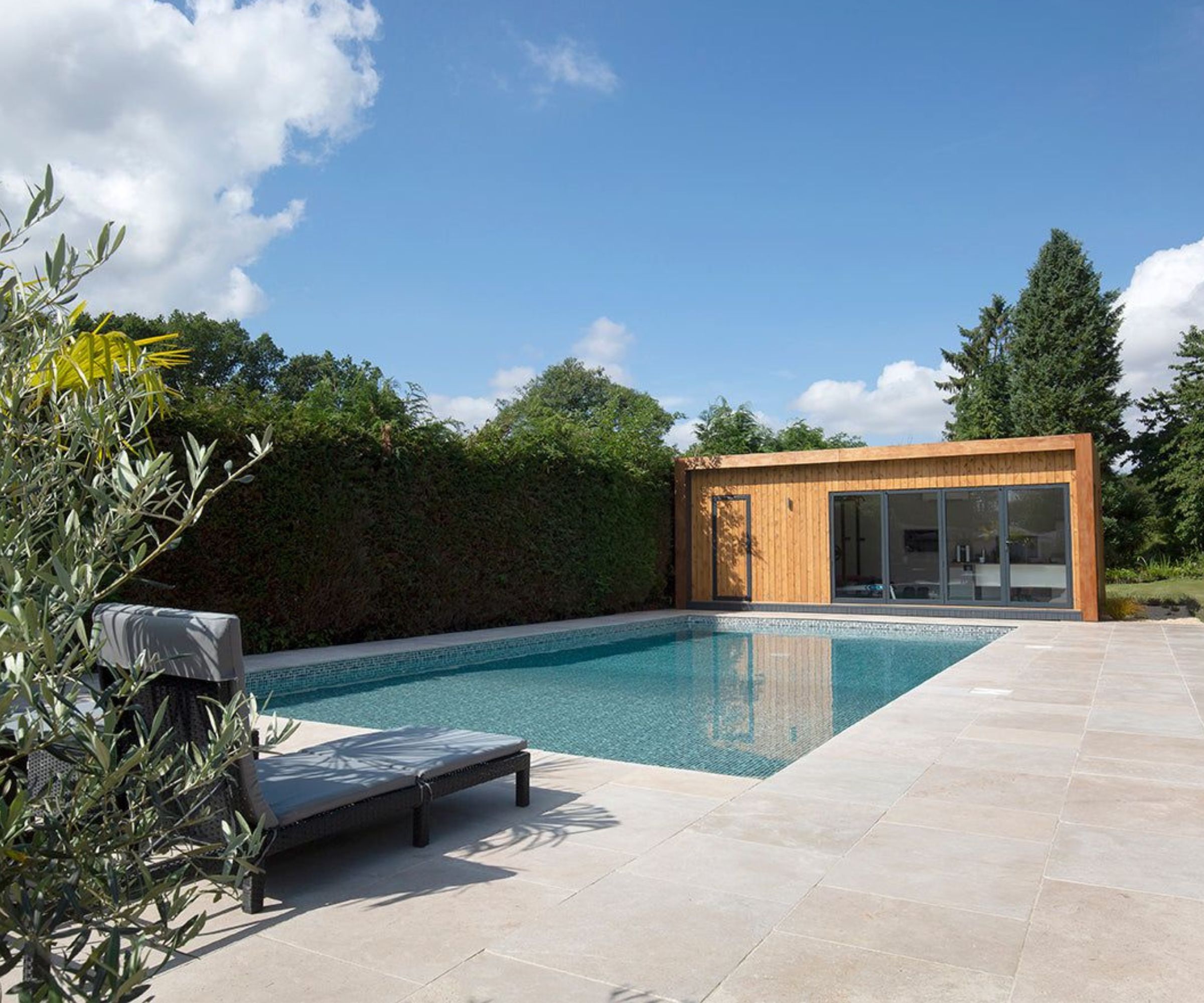 A rectangular outdoor pool with limestone surround, a grey sun lounger, olive tree and a cedar-clad flat-roof pool house with bifold doors and a yew hedge.