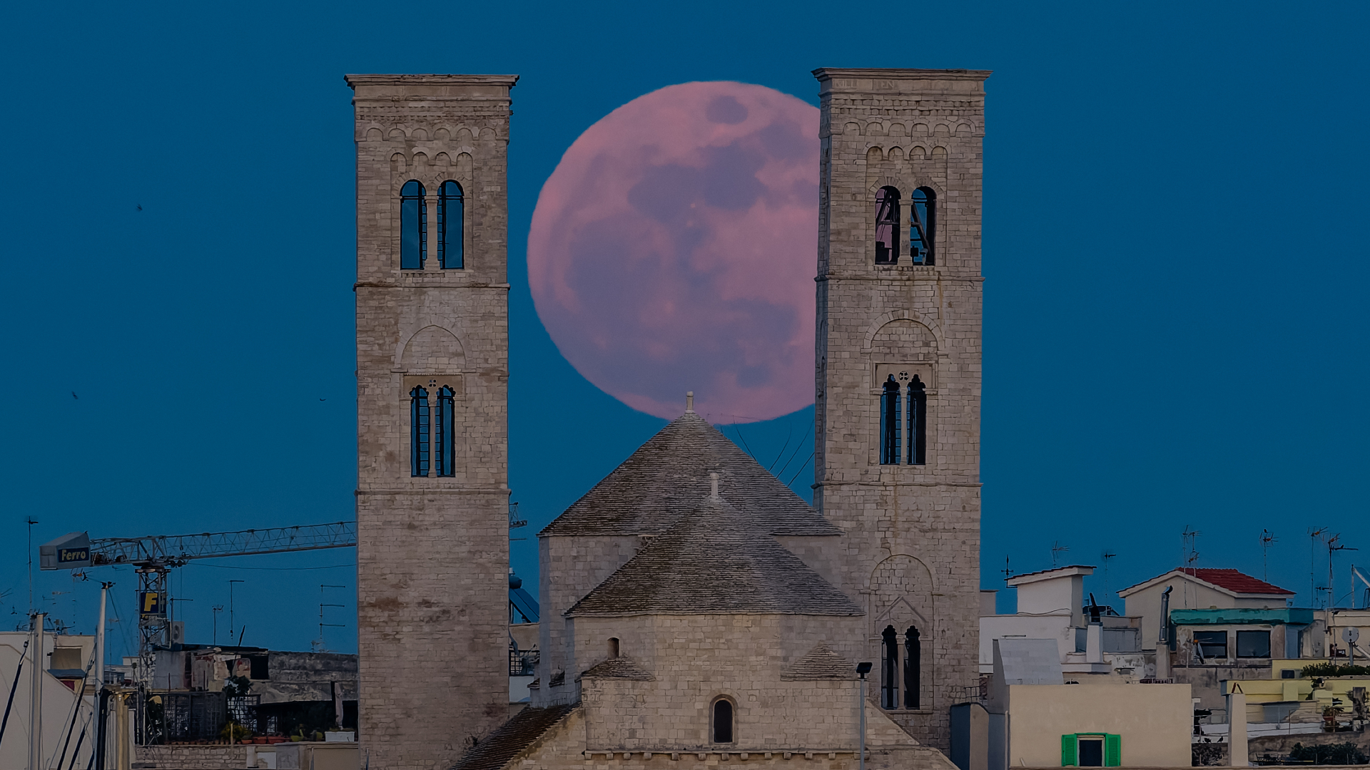 A red full moon rises in a dark blue sky between two stone towers that loom over the rooftops of a town in Italy.