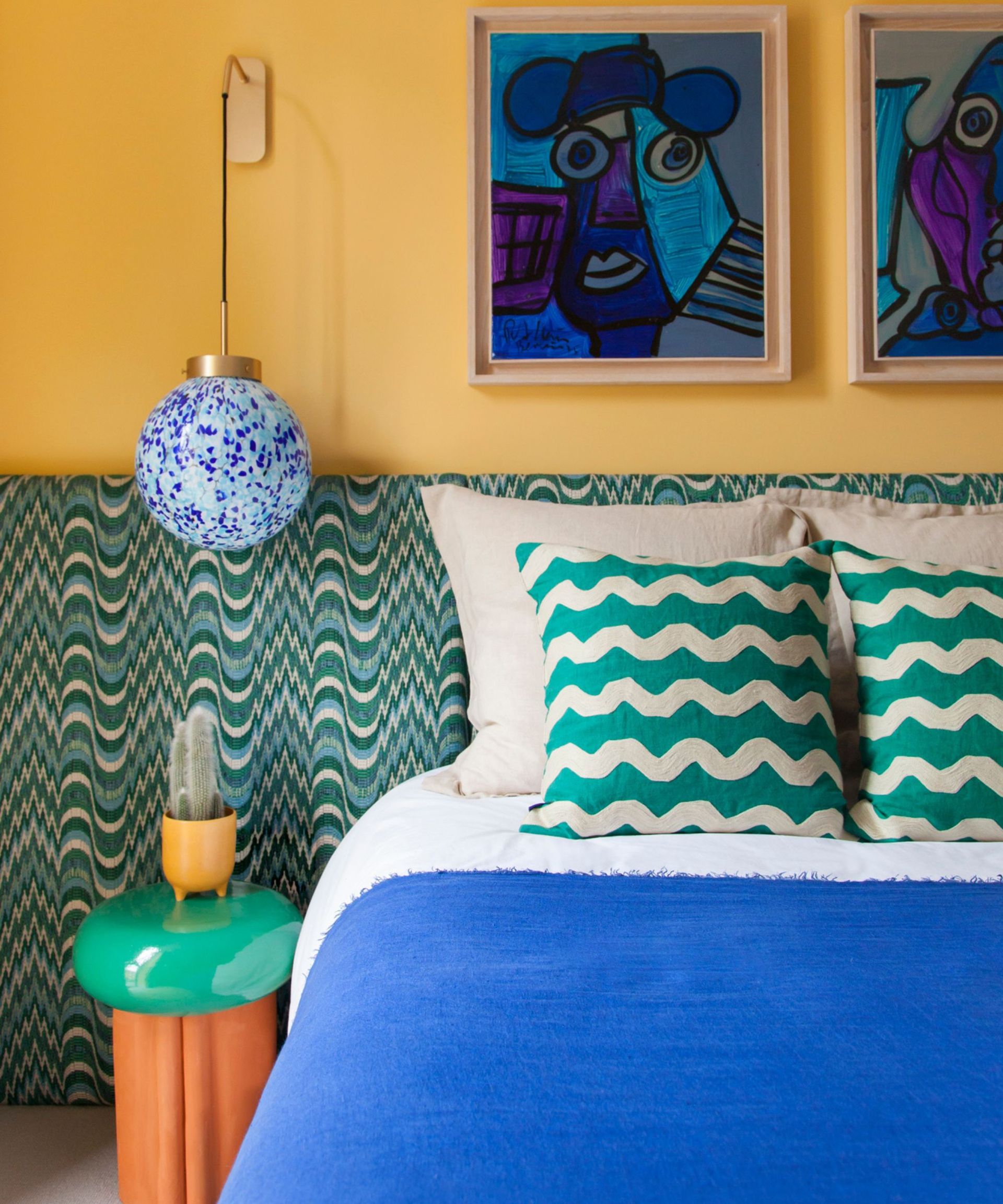 yellow painted bedroom with a wide green geometric headboard, a hanging pendant light and mid century abstract wall art in a bright blue