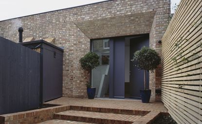 The front entrance of a brickhouse with staircase leading to the front grey door. Two potted mini trees on each side of the door 