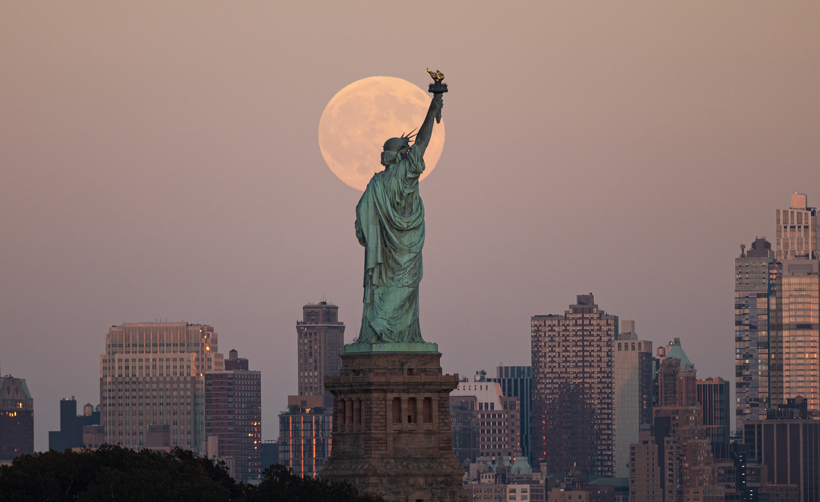A photograph of the Harvest supermoon behind the Statue of Liberty in New York City.