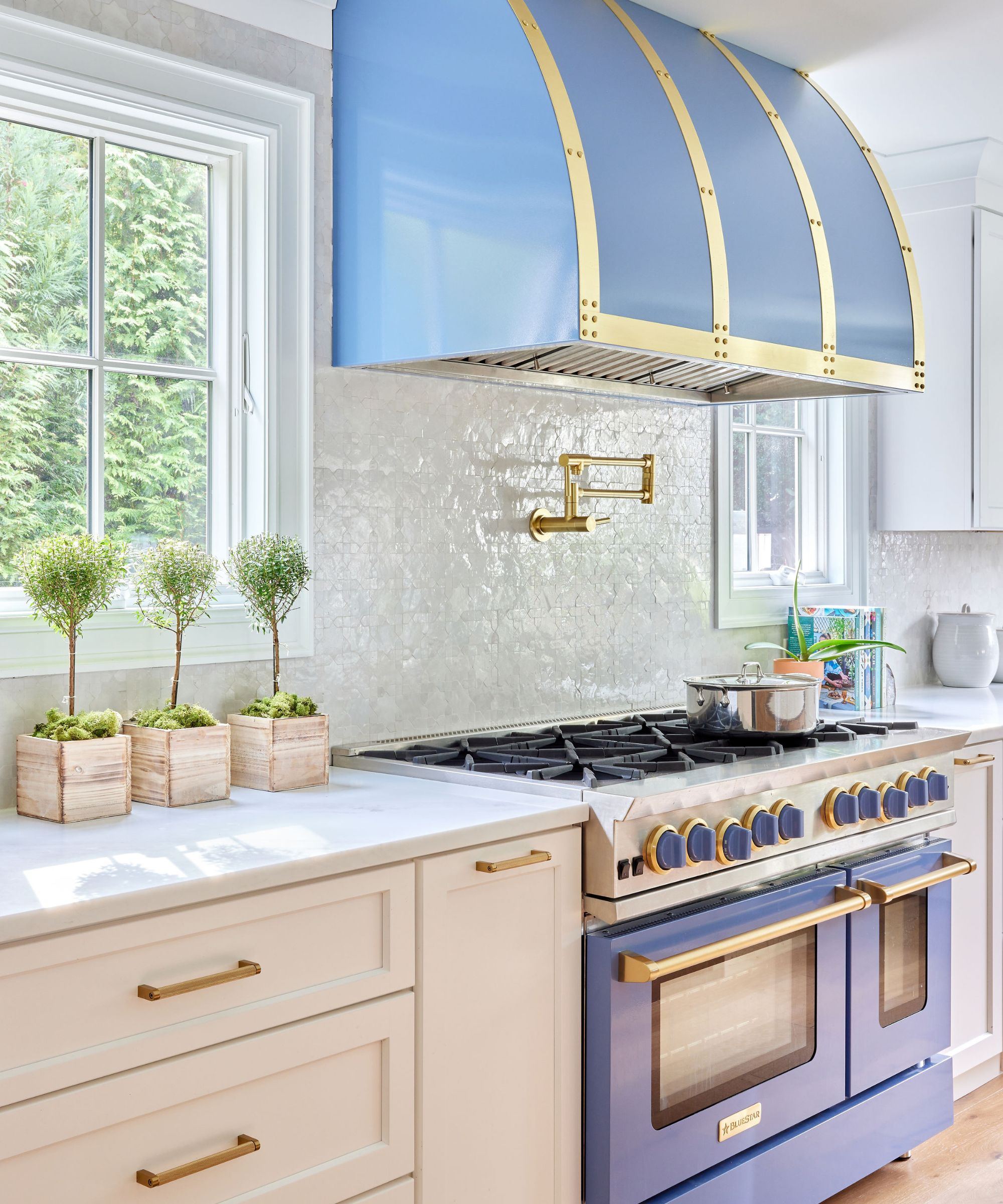 Kitchen with a cornflower blue range cooker and hood above, white tiles, a gold pot filler, white marble countertops, and white cabinets with gold handles.