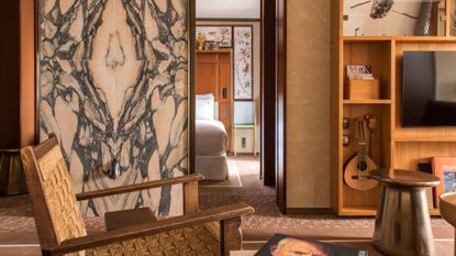 A hotel room with a relaxed rattan chair, a brushed copper stool, a carpeted floor leading to the bedroom and a wall unit with a tv, and a bandurria