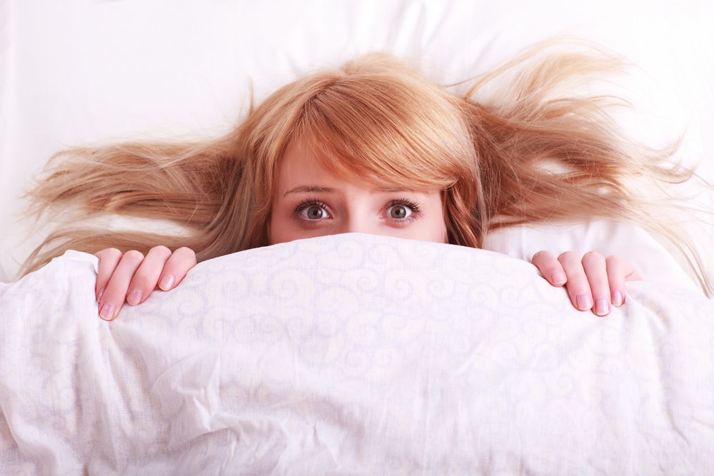 A woman lies in her bed, looking fearful.