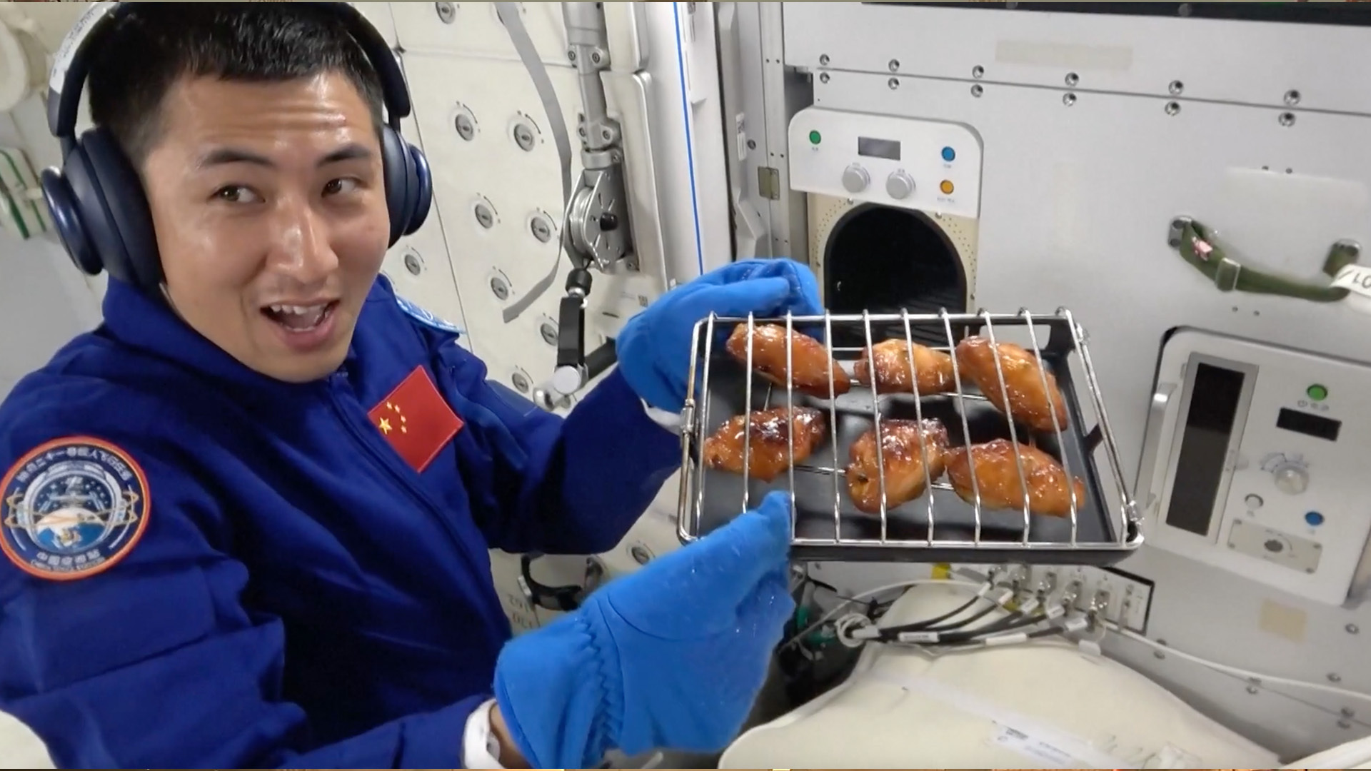 A man in a blue jumpsuit and mittens and headphones holds a grill tray with six chicken wing caged inside.