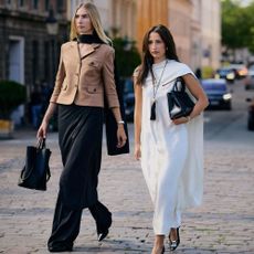 Stylish women pose in the streets of Copenhenhagen. The woman on the right wears a white dress with a cape and scarf elements with black accessories. The other woman wears a tan blazer, black scarf, black pencil skirt over trousers. 
