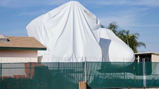 a large white cloth covers a full-size space shuttle model that's sitting between two buildings, behind a fence topped with razor wire