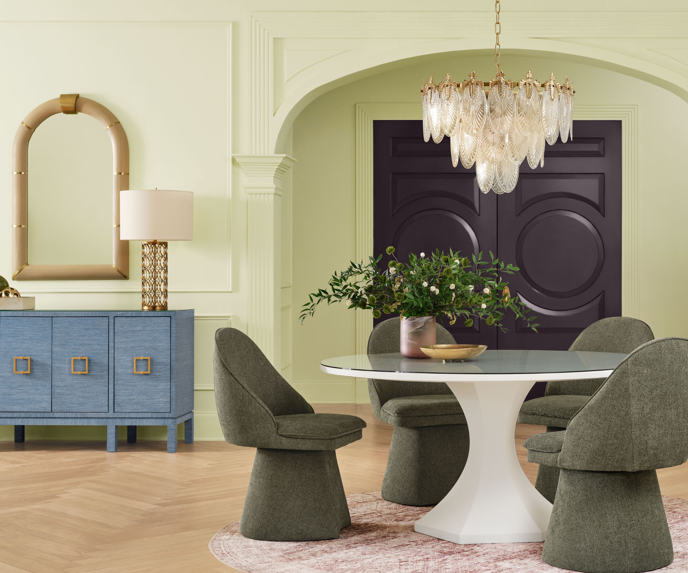 Dining room painted in Sherwin-Williams Celery, a dark aubergine front door, a round white table with dark green armchairs, and a blue console with a mirror and lamp.