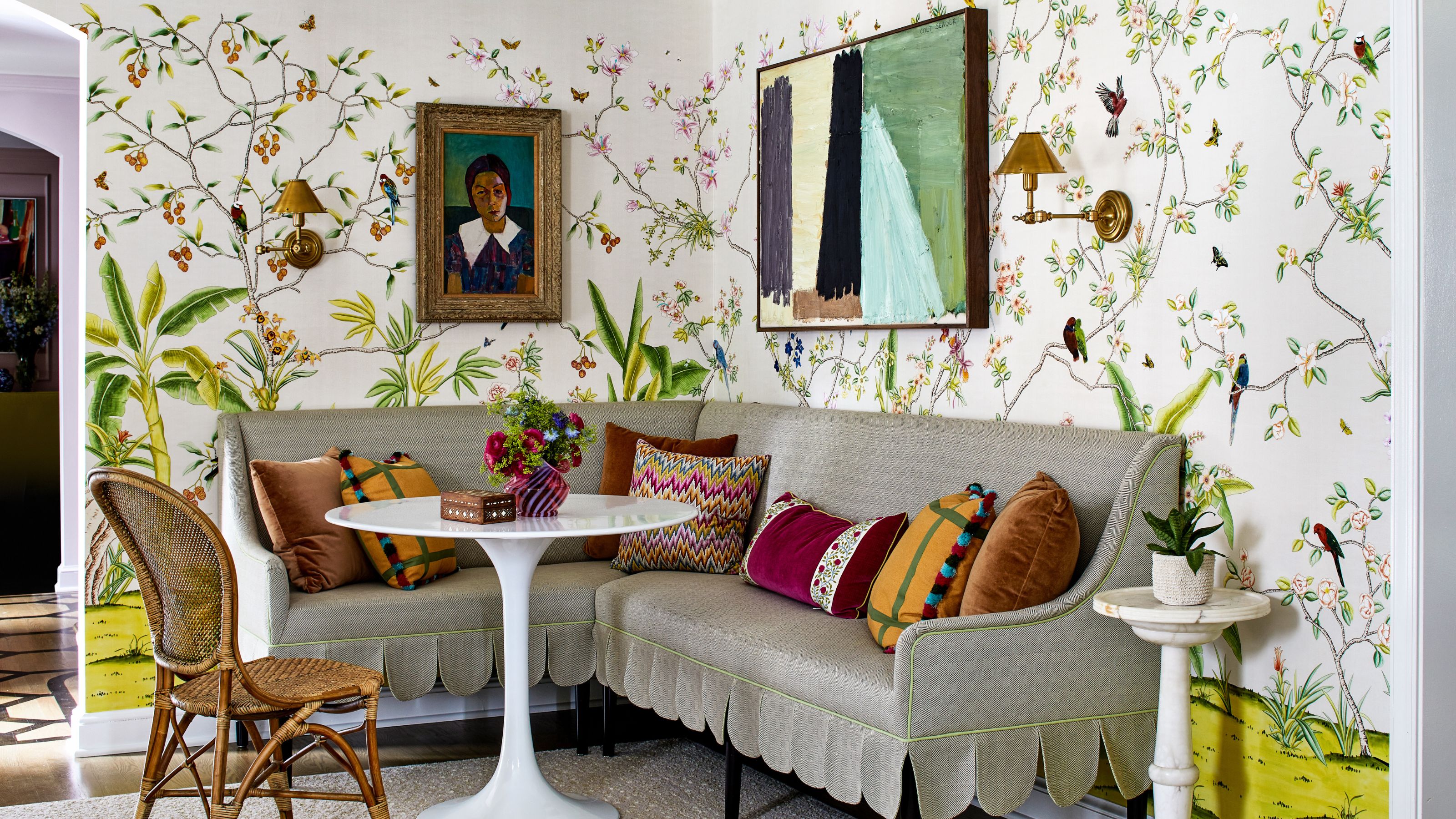 A seating space in the corner of a living room, featuring a striped corner banquette with scallop edges, a small white pedestal table, and throw pillows in various jewel tones