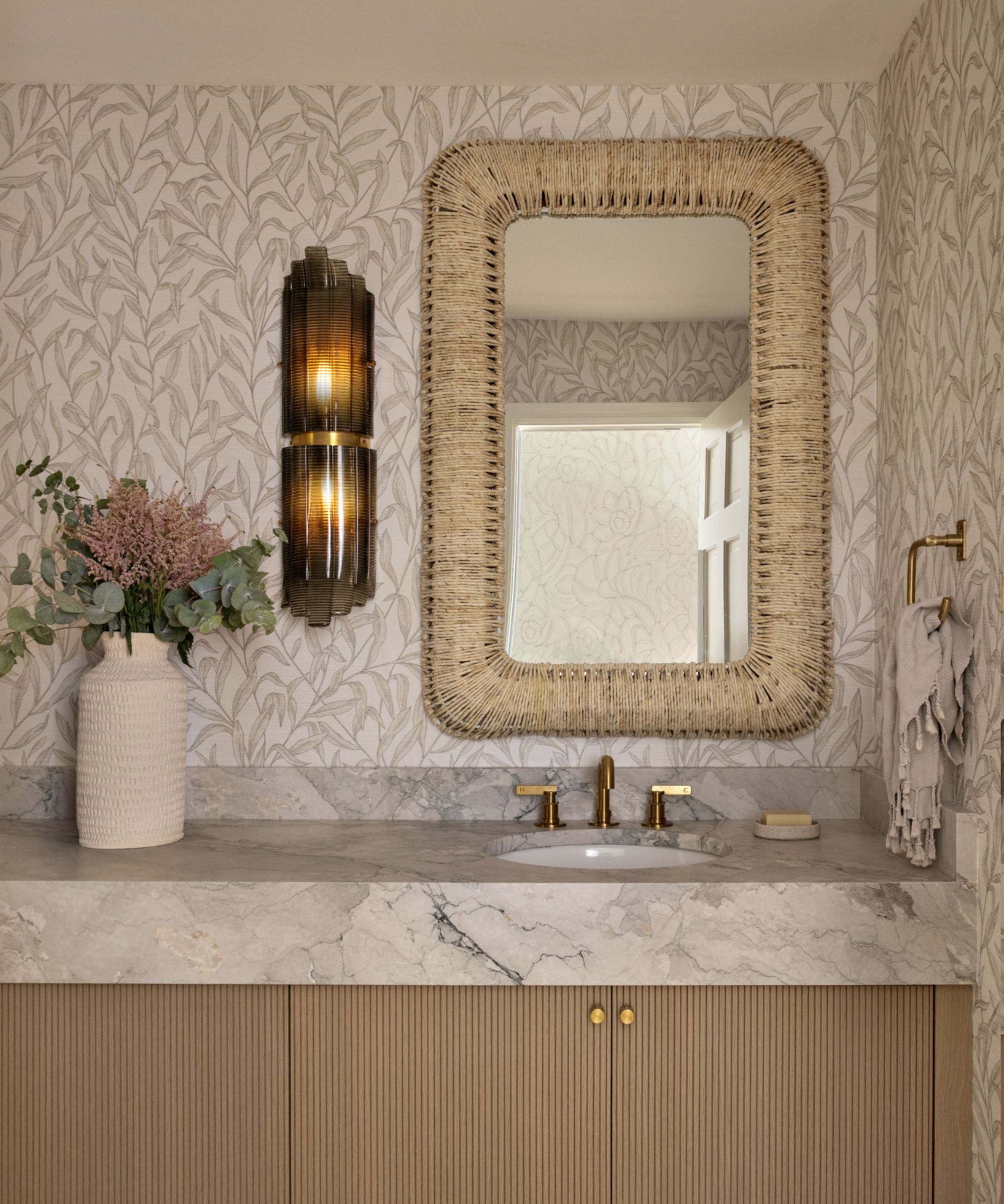 A neutral powder room with beige botanical wallpaper on the walls and a fluted wood vanity topped with a marble countertop