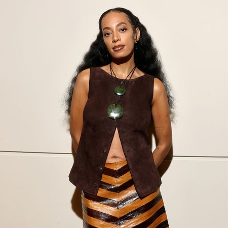 Solange Knowles (a woman with long, wavy hair) wearing a brown top and green necklaces looking at the camera.
