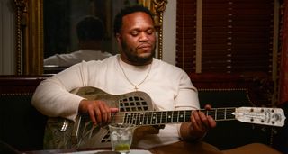 Jontavious Williams wears a white longsleeve and plays a cutaway resonator with a glass of water on the table in front of him.