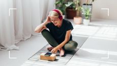 Woman sitting cross-legged on yoga mat with headphones on, browsing through the best Pilates apps
