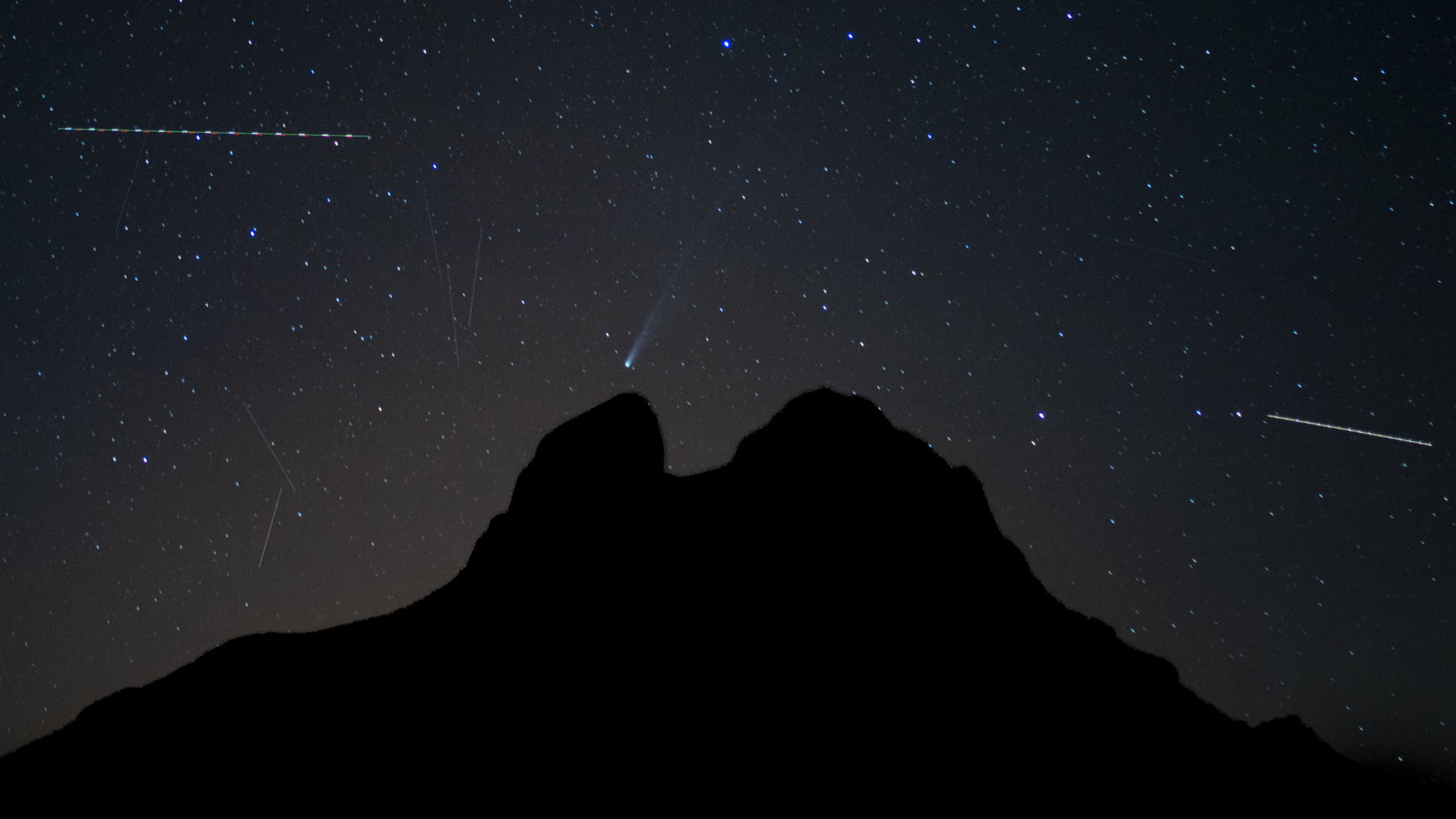 A comet is pictured streaking down towards a silhouetted mountain through a starry sky filled with satellite streaks.
