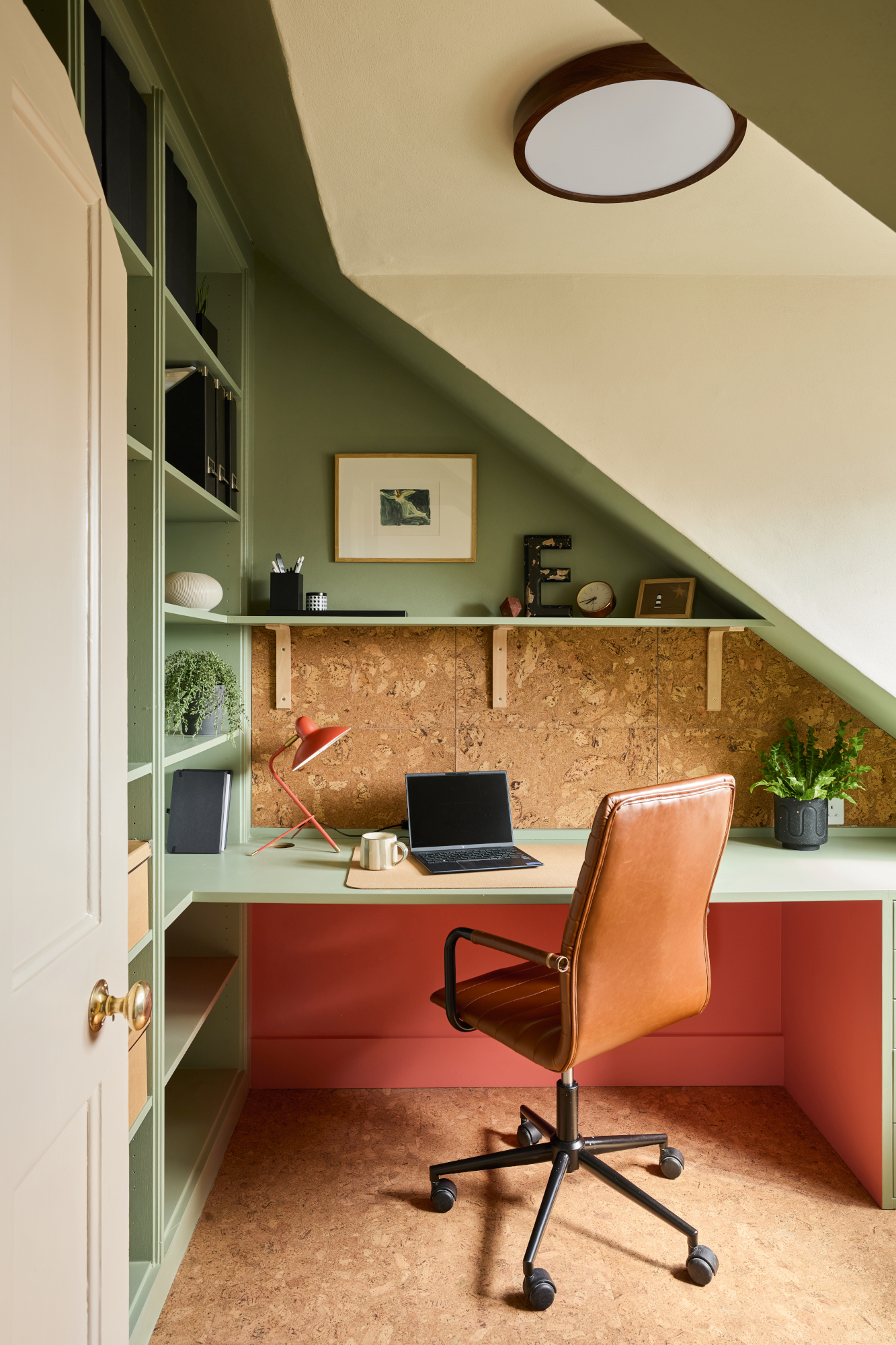 Home office tucked in the eaves with green walls and shelving, cork wall and floor, tan desk chair and coral underside