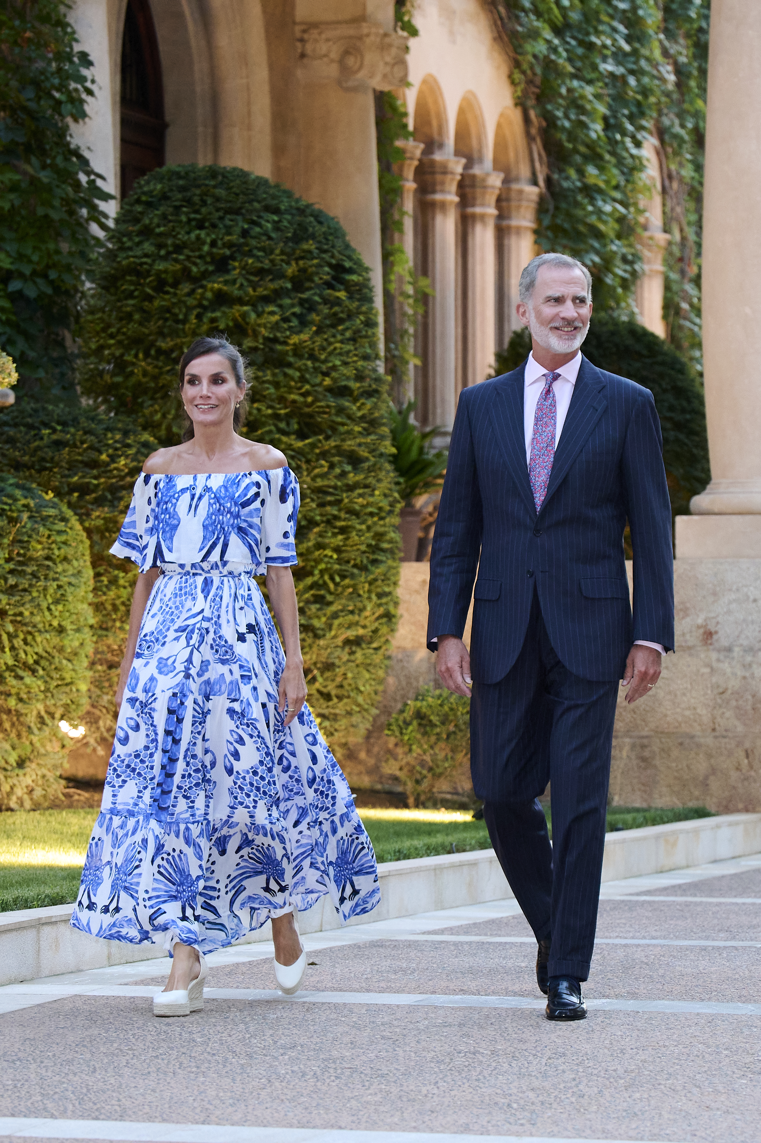Queen Letizia wearing a blue and white dress walking next to King Felipe outside columns and bushes