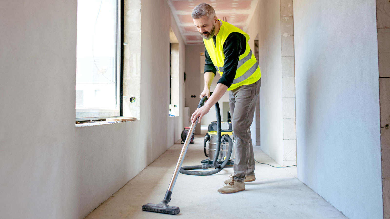 Man in hi-viz yellow vest using a wet and dry vacuum in hallway with bare plastered walls