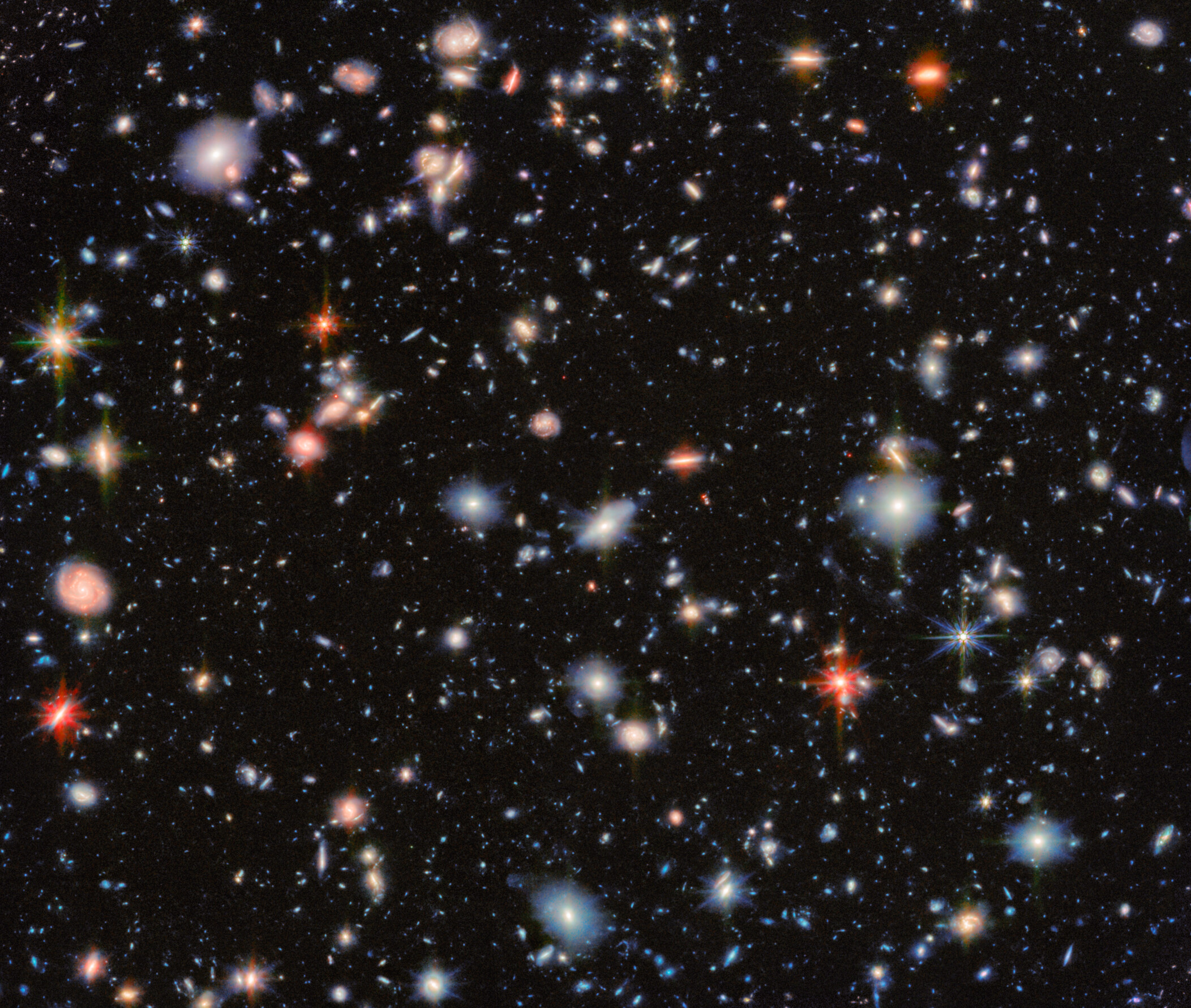 A photo of galaxies and stars in deep space, with glows of red and blue and orange stars with swirls of small galaxies.
