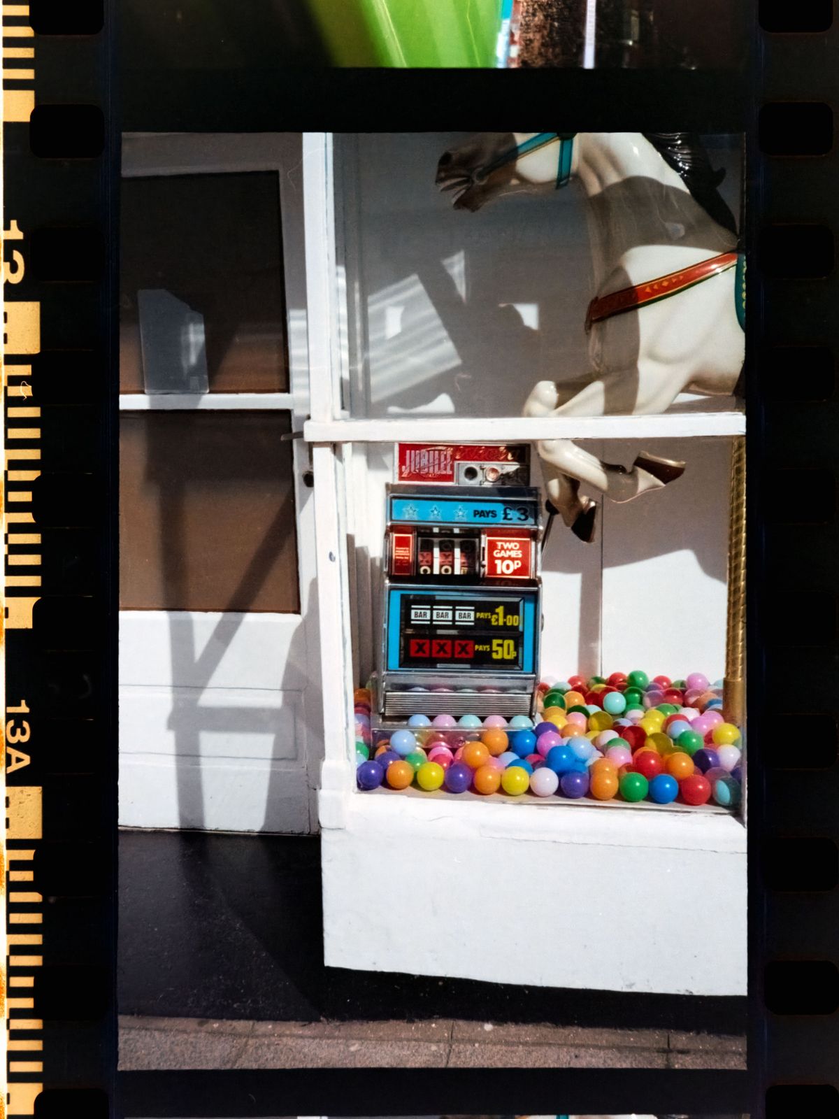Scanned color negative showing a seaside amusement arcade window with a vintage slot machine, colorful plastic balls, and part of a carousel horse.