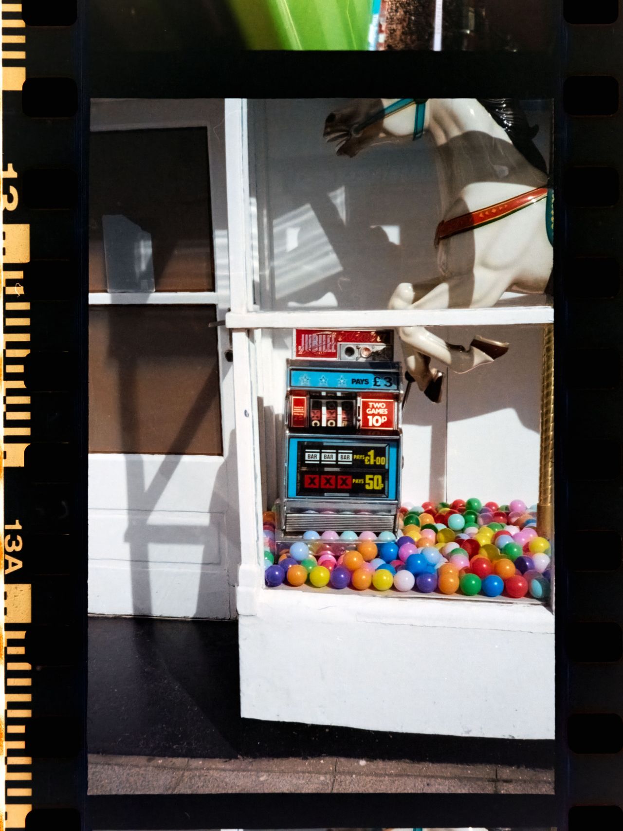 Scanned color negative showing a seaside amusement arcade window with a vintage slot machine, colorful plastic balls, and part of a carousel horse.
