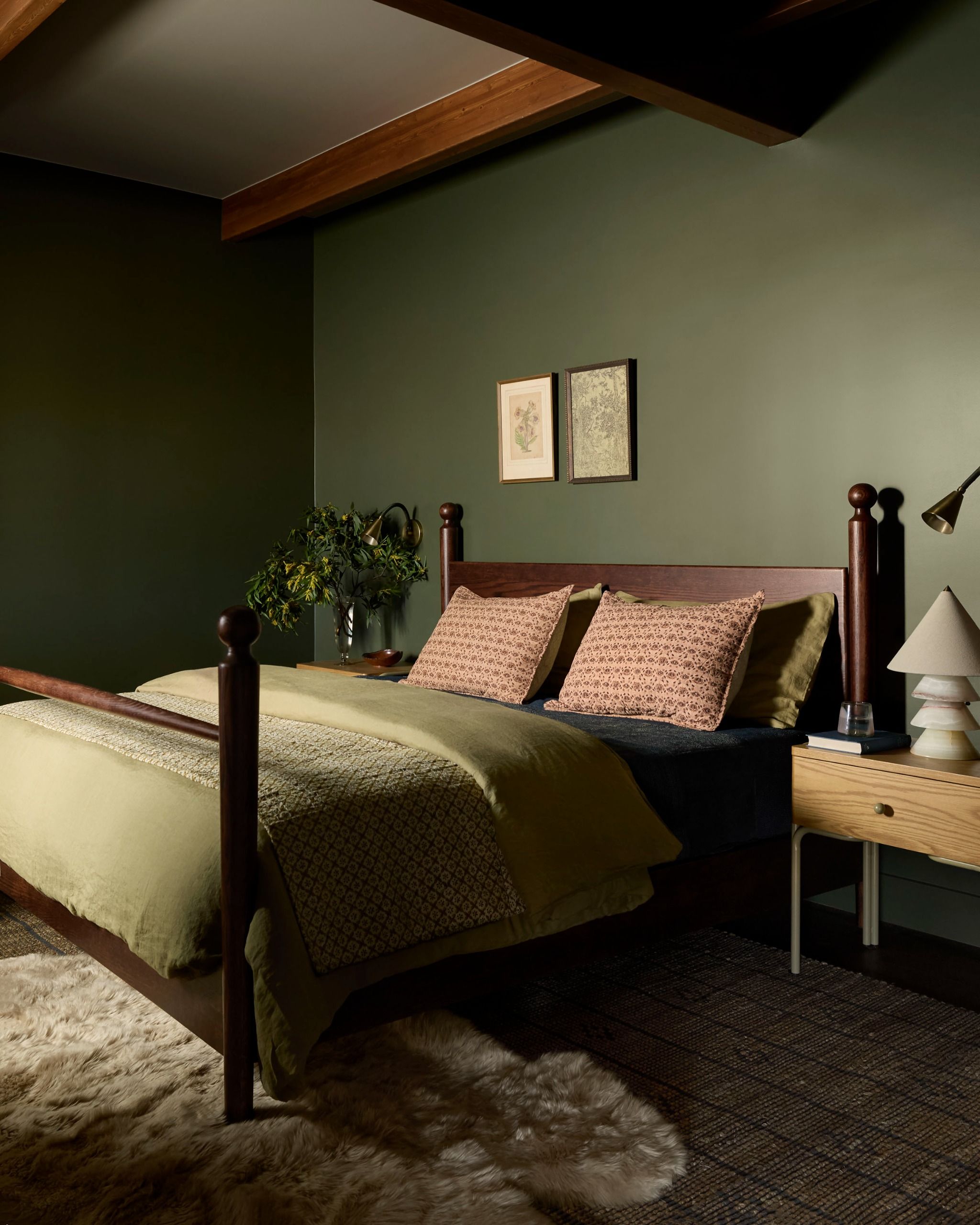A modern bedroom with dark green walls, green bedding with pink cushions and a cream rug by the foot of the bed, artwork to the left of the bed, and a vase of flowers on one side and a lamp the other