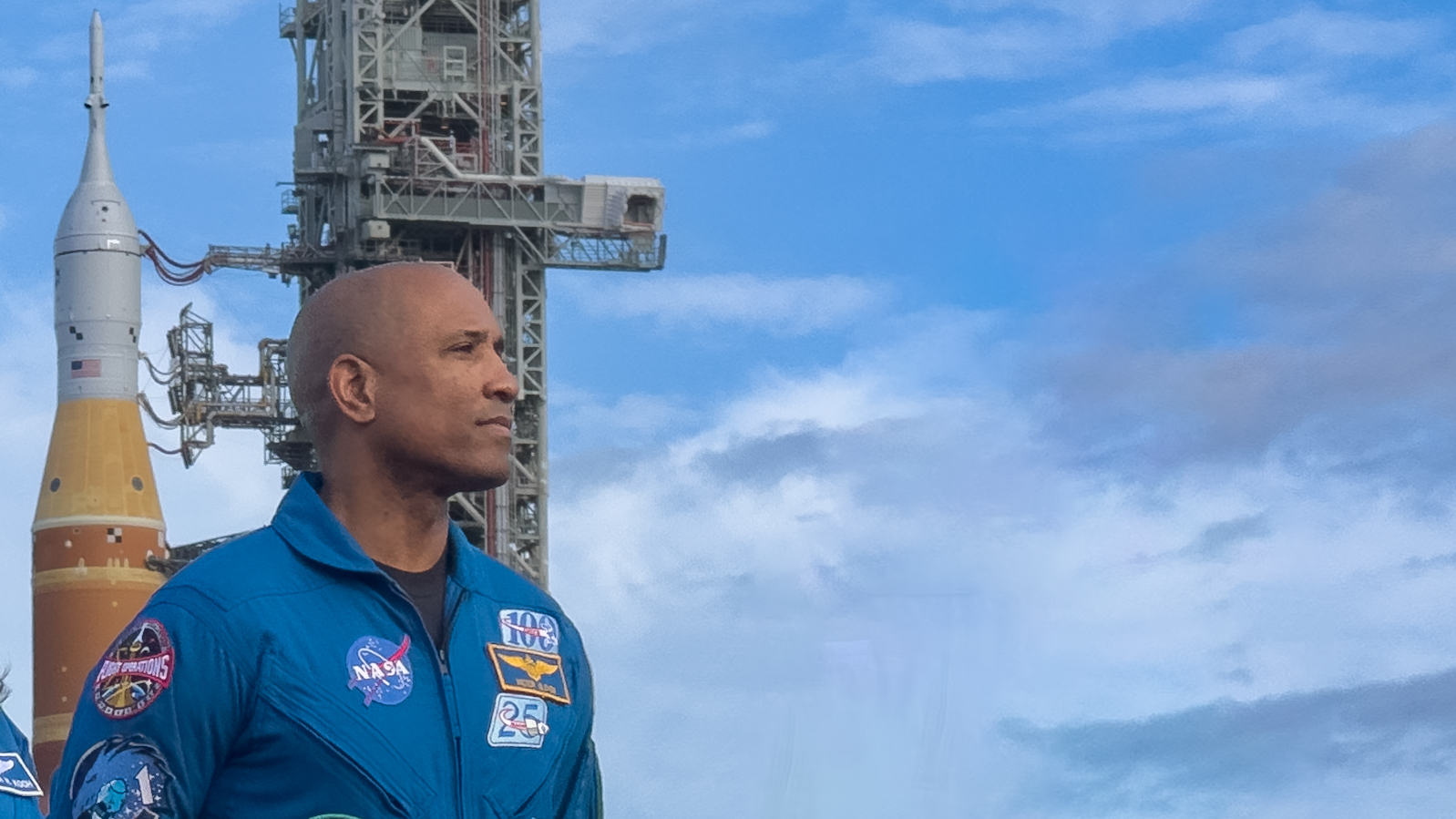 A man in a blue jumpsuit stands against a blue sky with a big orange rocket behind him on the left.