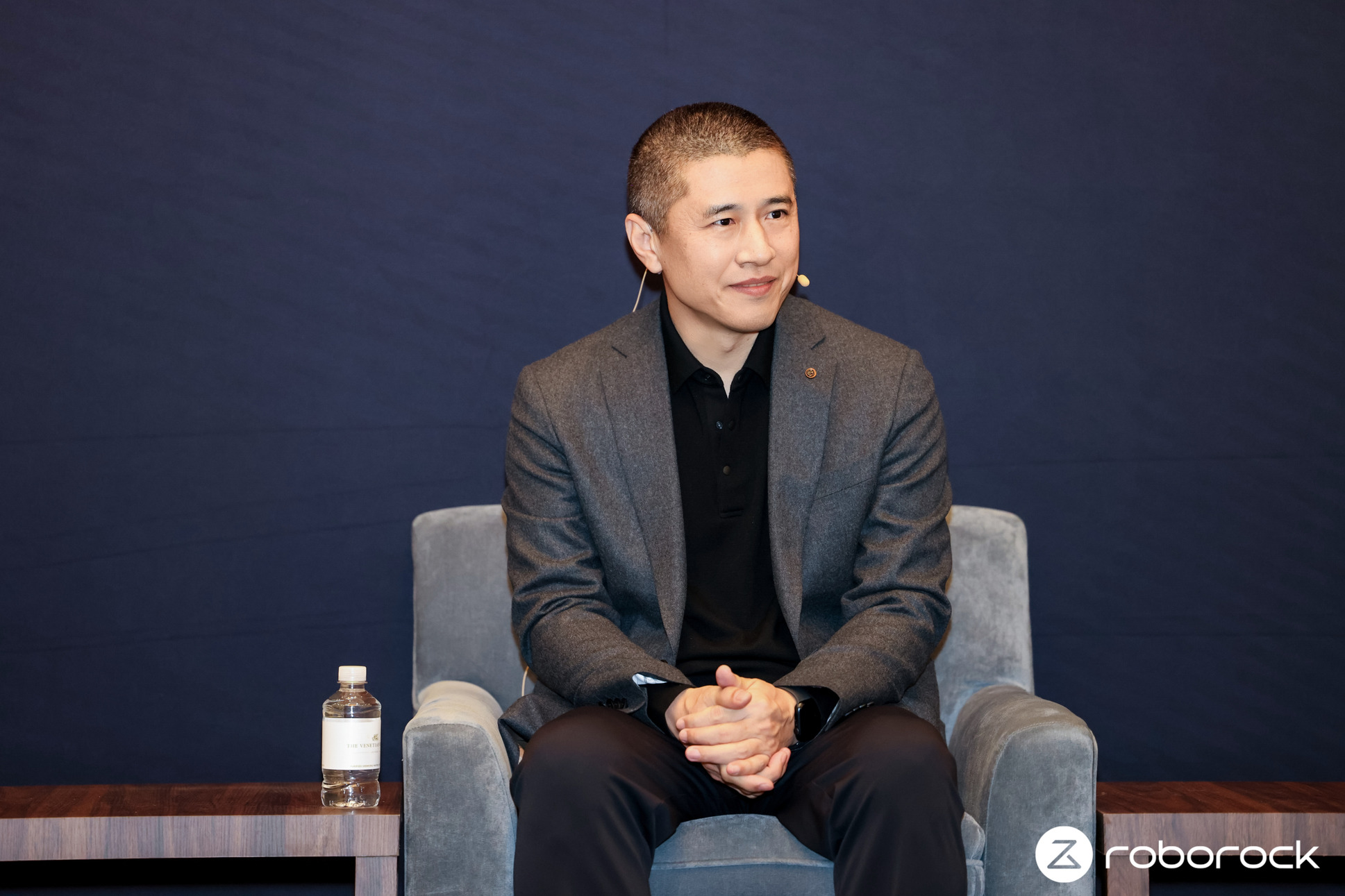 Photograph of Quan Gang sat on gray armchair wearing dark gray suit, smiling and looking attentively to his left.