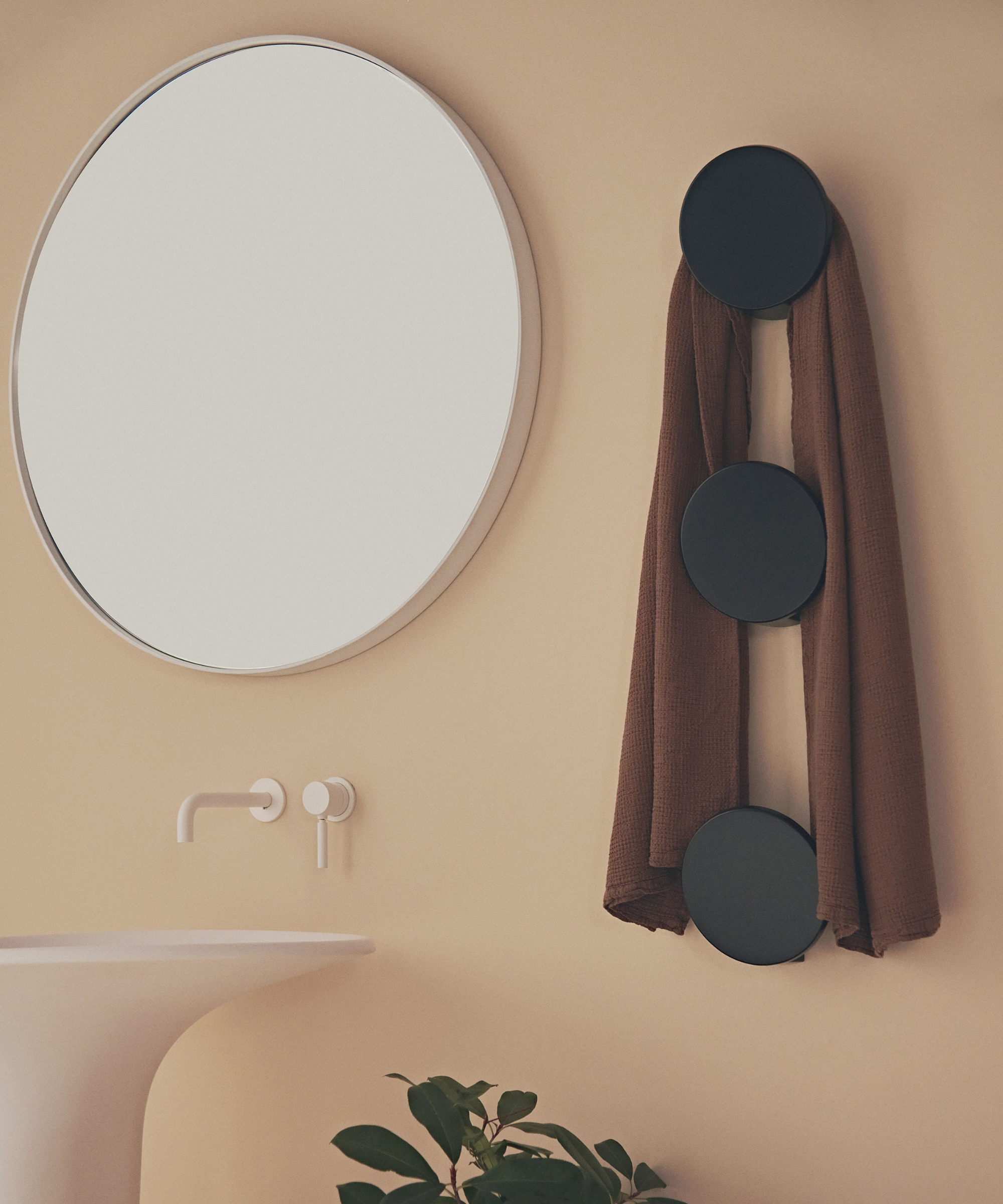 A warm sand-toned bathroom wall featuring a large round mirror with a thin white frame above a wall-mounted white tap and spout. To the right, three circular matte black disc-shaped hooks are mounted vertically, holding a draped terracotta linen towel.