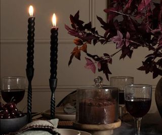 Two twisted black taper candles and glasses of dark wine, next to a dark plant.