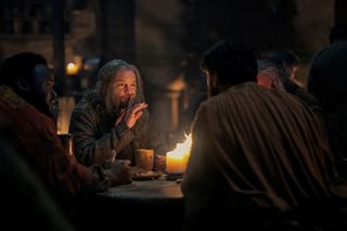 Tom Bennett dans le rôle d'Ulf le Blanc (au centre), assis avec d'autres hommes à une table tenant des bougies, dans « La Maison du Dragon »