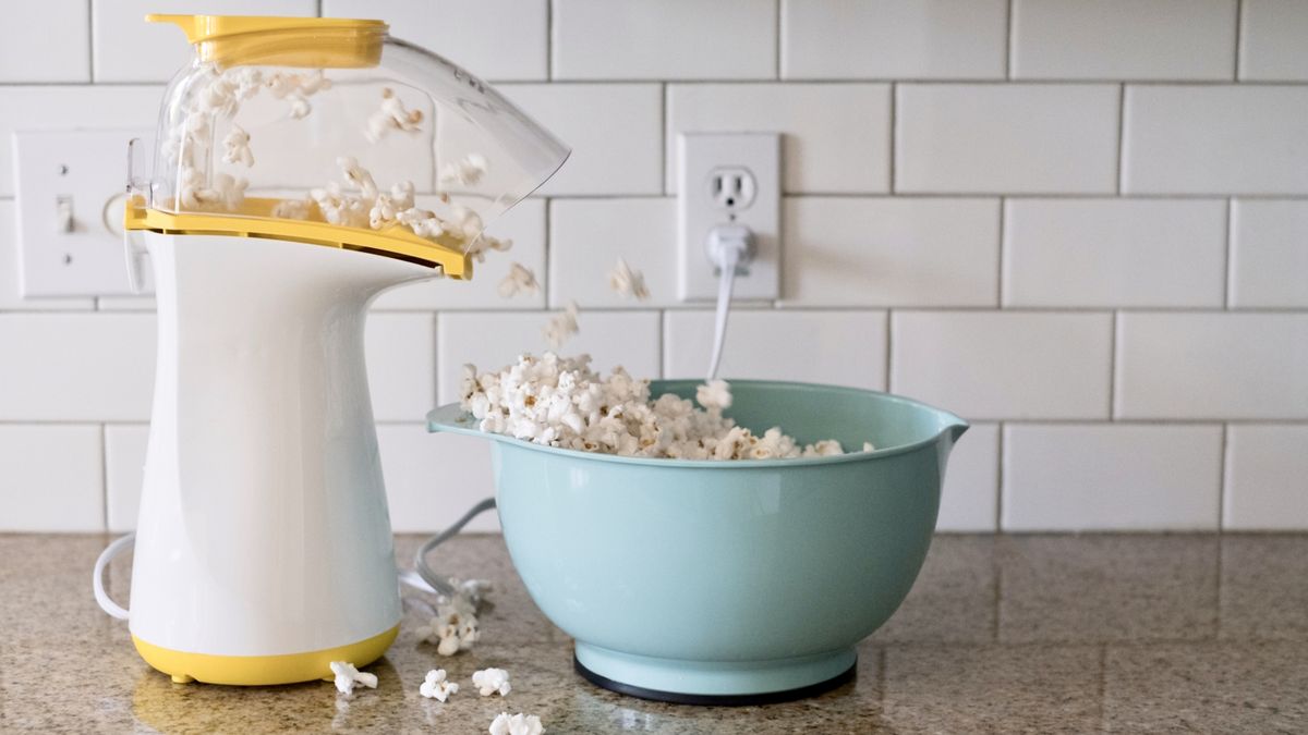 Popcorn machine popping popcorn into a aqua colored bowl on the countertop of a residential kitchen. The popcorn is overflowing onto the counter.