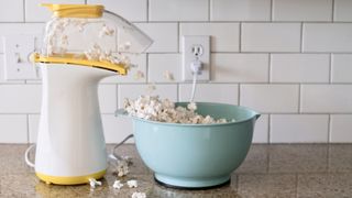 Popcorn machine popping popcorn into a aqua colored bowl on the countertop of a residential kitchen. The popcorn is overflowing onto the counter.