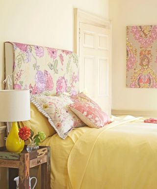 A yellow bedroom with a purple florall wall mounted headboard, a yellow bed with throw pillows, and a wooden side table next to it with flowers and a yellow lamp next to it